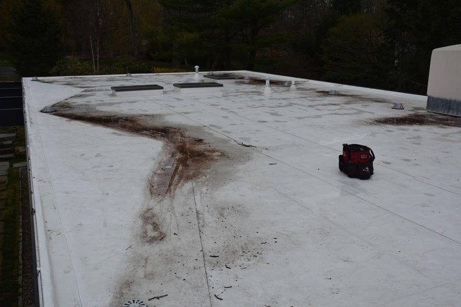 Dirty white TPO flat roof with visible stains, debris, and a red tool bag placed on the surface