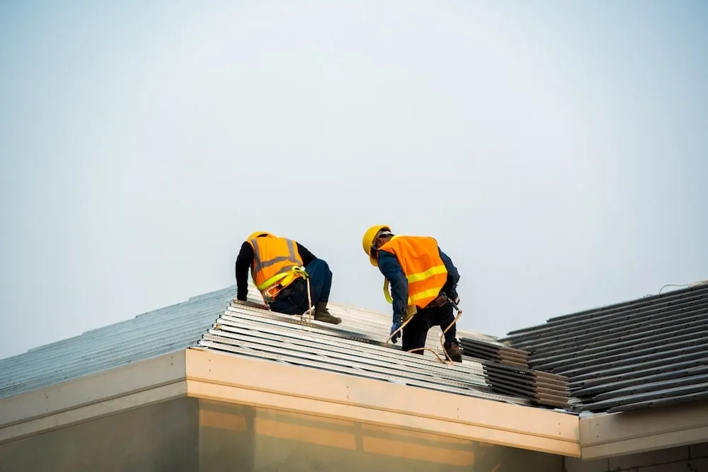 Two construction workers in safety gear installing roofing tiles on a house