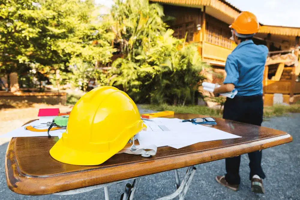 Yellow hard hat on a table with blueprints at an outdoor construction site.