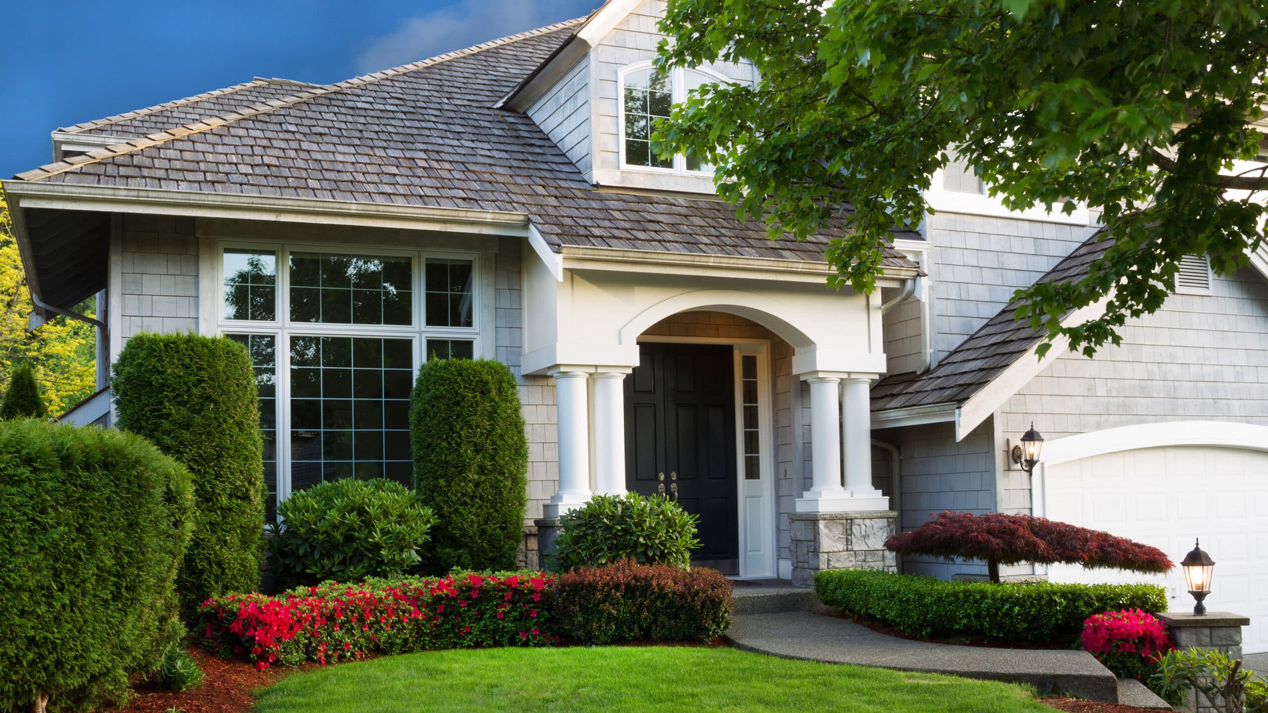 Elegant suburban home with gray siding, large front windows, black door, manicured bushes, and vibrant garden landscaping
