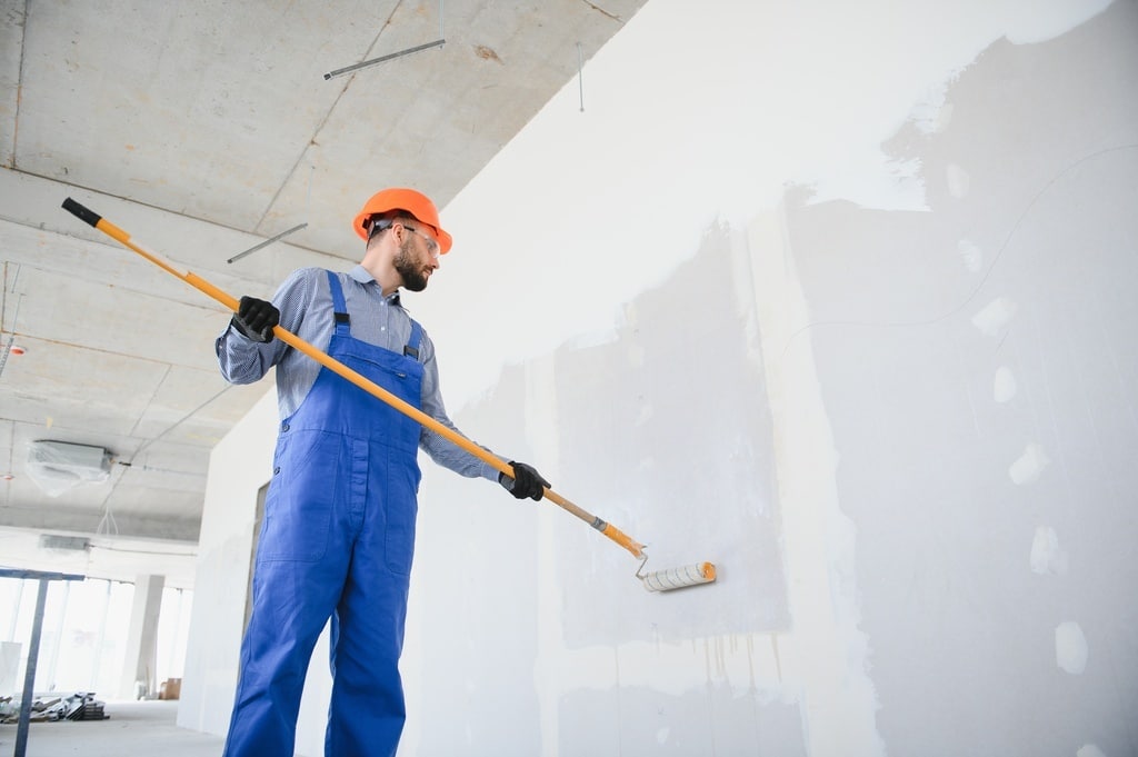 A man in blue overalls and a safety helmet is painting a wall with a roller on a construction site