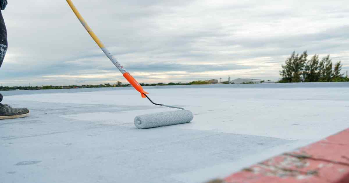A person painting a rooftop with a roller, applying a coat of white waterproofing paint