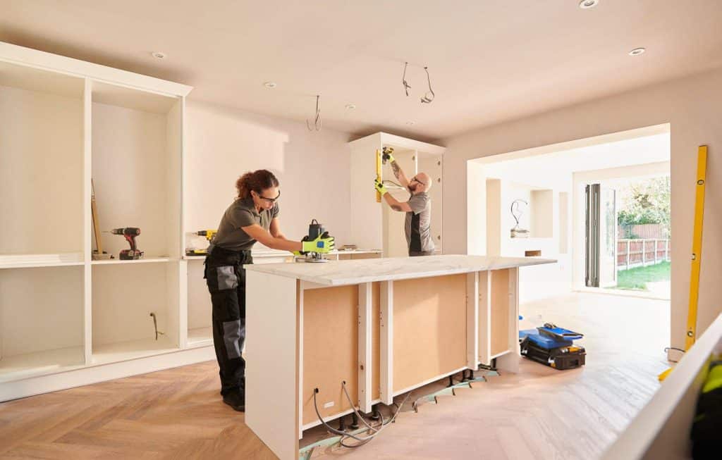 Two workers are renovating a kitchen, one working on the countertop and the other installing a cabinet